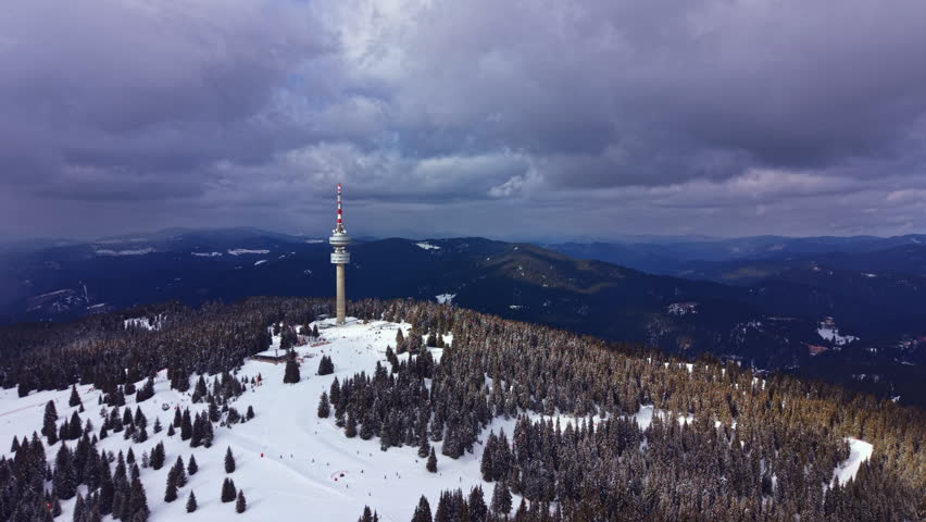 A tall communication tower stands on a snowy mountain. Trees are covered in snow, and hills can be seen in the background. Dark clouds hang overhead in winter.