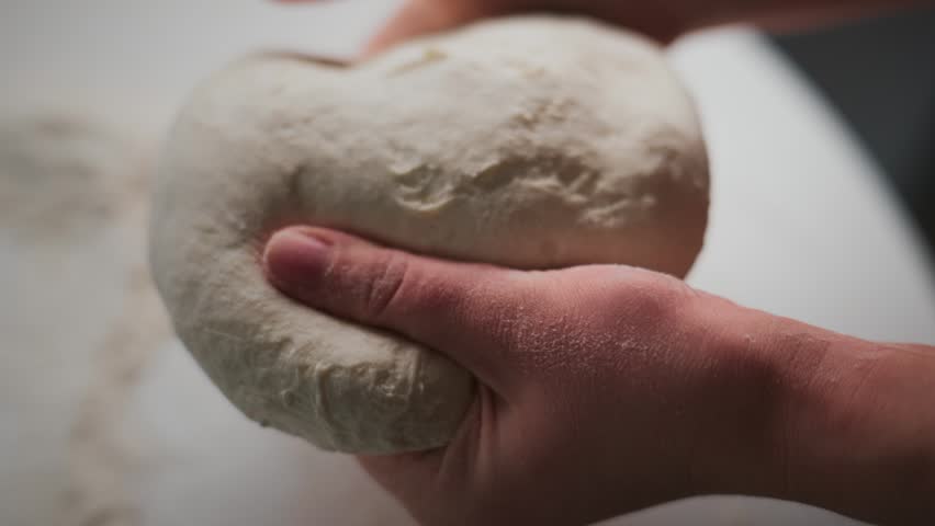 Hands gently knead fresh dough on a floured surface. Fingers press into soft, elastic dough. Texture shows rise and moisture of homemade bread. Close-up focuses on tactile process of dough preparation