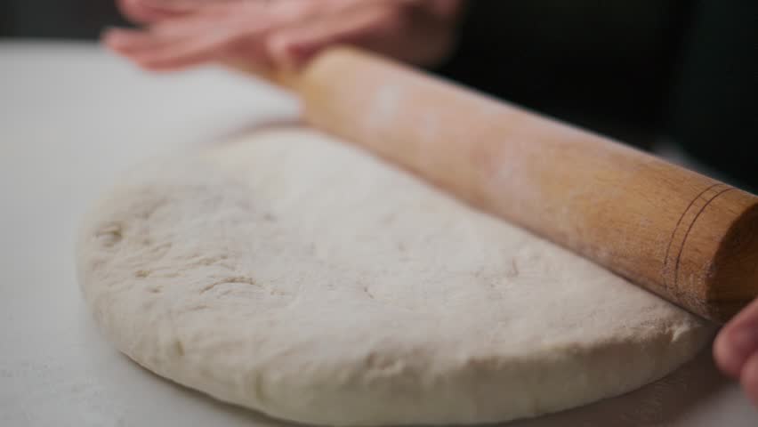 Person rolls dough with a wooden roller on a white surface. Hands apply gentle pressure as the roller flattens the dough. The focus stays on the motion and texture of the dough