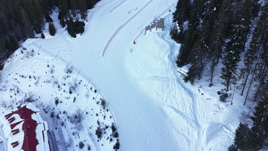 Snow covers the ski slope as people enjoy skiing and snowboarding. Trees line the edges of the trail, and a lodge is seen nearby. The sky is clear and bright above the mountain.