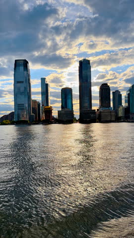 Sunset behind Jersey City skyline from the Hudson River, New York. Golden light through clouds, glass towers silhouetted, and rippled water reflections.