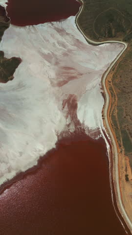 Vertical aerial view of red water, white mineral deposits and textured shoreline at an industrial pond.