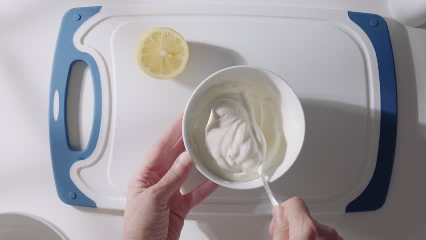 Hands stir Greek yogurt in a bowl while preparing a creamy homemade sauce with fresh ingredients