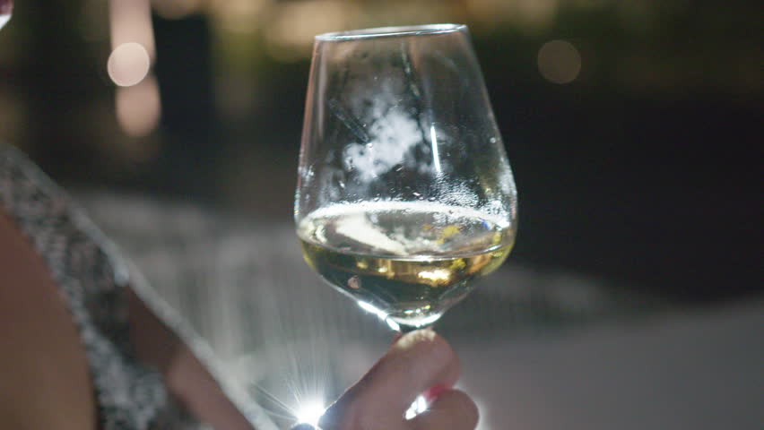 Close-up of a senior woman holding and drinking crisp white wine from a glass at night. The backlight highlights her hand and the condensation on the cool beverage. She sips the drink.