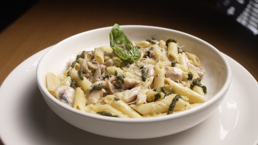 Freshly made penne pasta with chicken, mushrooms, and pesto in a white bowl rotating slowly, fa01