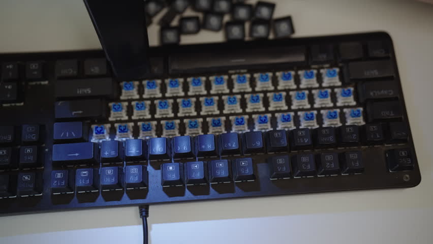 Close-up view of technician hand using portable vacuum cleaner to remove dust and debris from switches of dirty mechanical keyboard during regular maintenance and cleaning. Shooting in slow motion