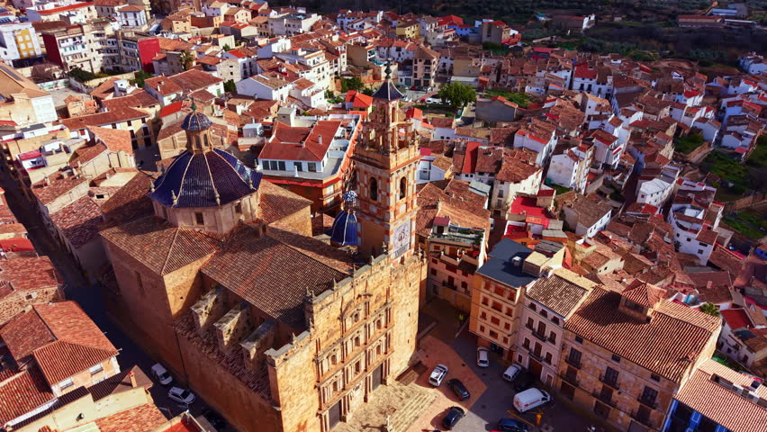 The scene captures a high view of a village with many houses and buildings. Roofs are mostly red. Two towers stand out against the skyline. A few cars are parked on the road below.