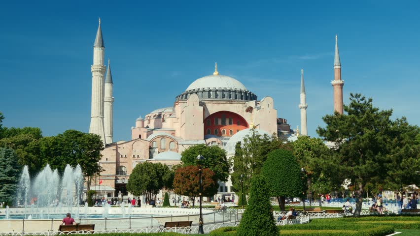 Historic Hagia Sophia Grand Mosque with a decorative water fountain spraying in Sultanahmet Square park under a bright blue sky in Istanbul, Turkey. Wide static shot 