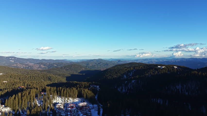 Mountain landscape shows snow on trees and hills under a clear blue sky. Distant peaks and valleys can be seen. This scene captures winter nature in a broad view.