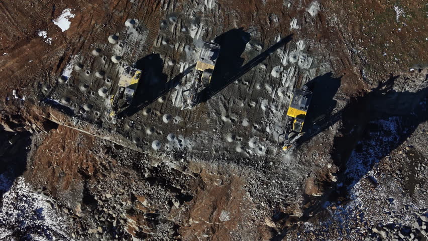 Machines work on the surface of the granite quarry. The ground is marked and the area is surrounded by rocks. This is happening during daylight hours in an open space.