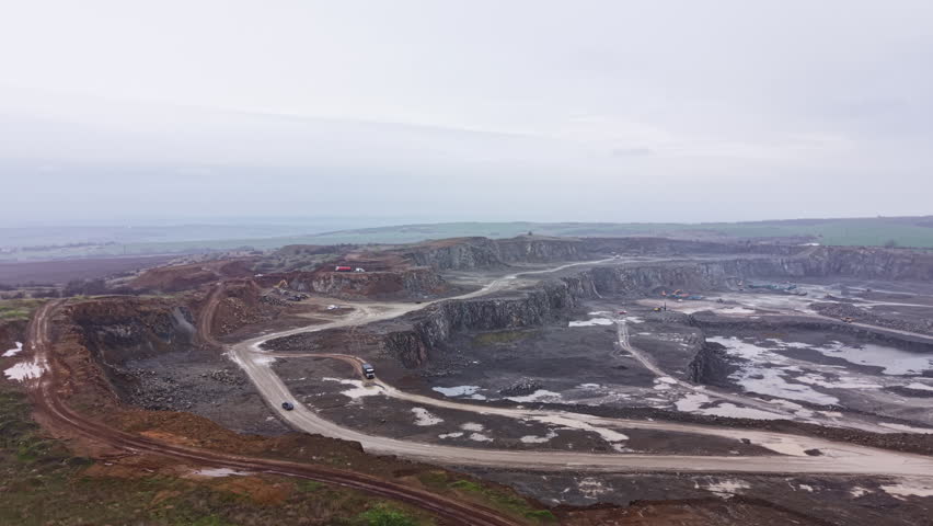 A granite quarry is visible with large sections of rock cut away. Heavy machinery operates on the site. Dust and mud cover the roads. The setting shows a cloudy sky over the open landscape.