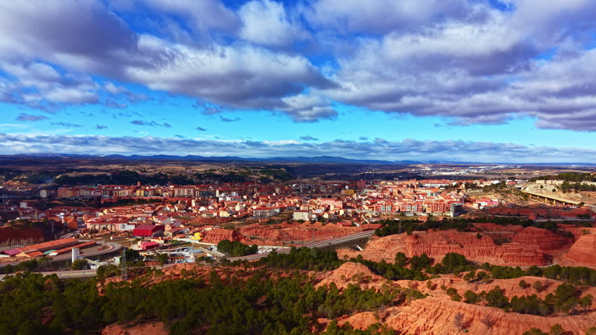 The scene shows a large town with a mix of buildings and roads. Red hills are visible in the foreground under a blue sky with clouds.