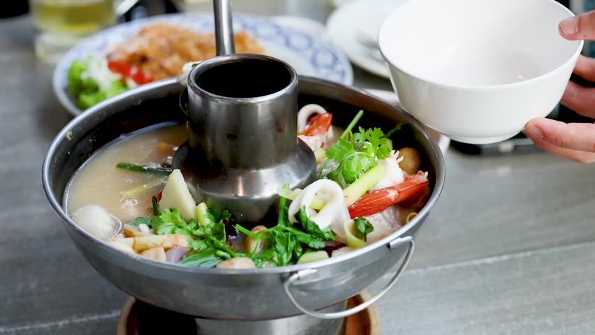Close-up shot of hands ladling spicy seafood soup from a traditional metal hot pot