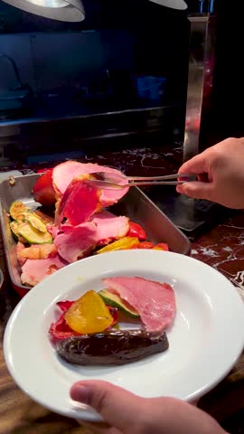 A person uses metal tongs to serve sliced roasted ham onto a white plate