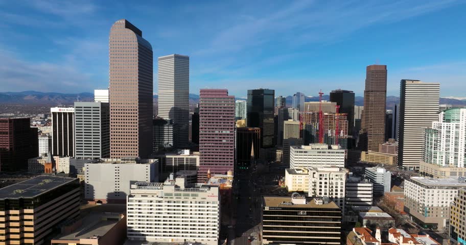Aerial drone shot panning forward slowly, showcasing downtown Denver, Colorado skyline with its majestic buildings under sunny, blue sky, and distant Rocky Mountains in the background, daytime footage.