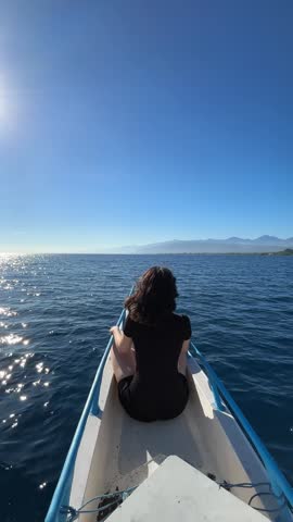 Woman sitting on bow of speeding boat in blue ocean near Bali coastline.