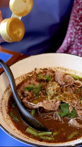 A close-up shot shows fish sauce being poured from a bottle into savory pork noodles.