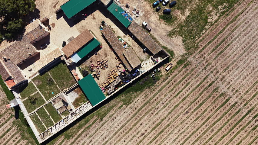 Aerial view shows farm buildings arranged around a central area with several vehicles and parked cars in a rural landscape under clear skies.