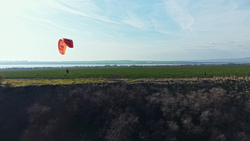 Two individuals walk near the edge of a hill while a paraglider soars above green fields and a lake in the distance under a clear sky.