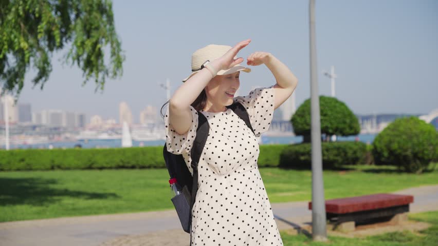 A smiling young female tourist poses happily in a sunny waterfront park, wearing a polka dot dress, straw hat and backpack, enjoying her leisure travel time.