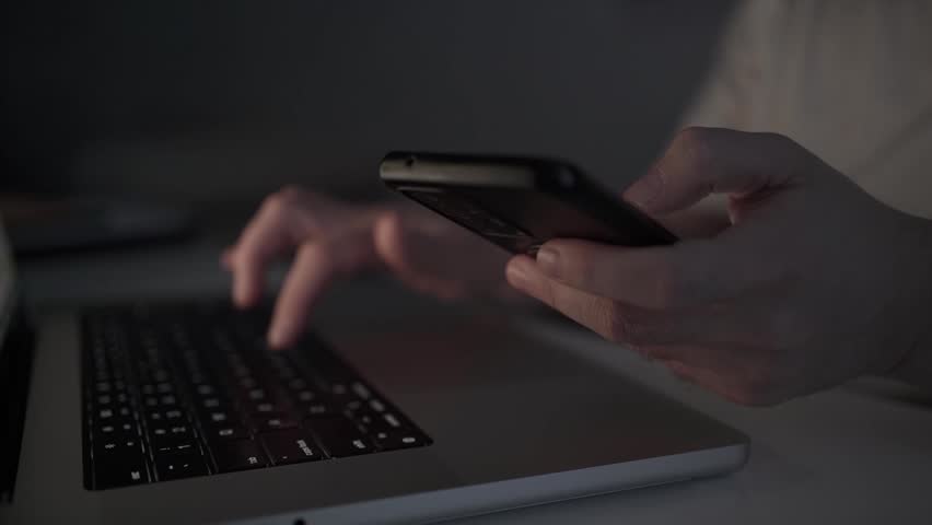 Man using a smartphone while working on a laptop at a desk. Modern multitasking, digital communication, remote work, productivity, and technology concept.