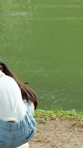 A young girl in denim shorts crouches by a green pond in natural daylight