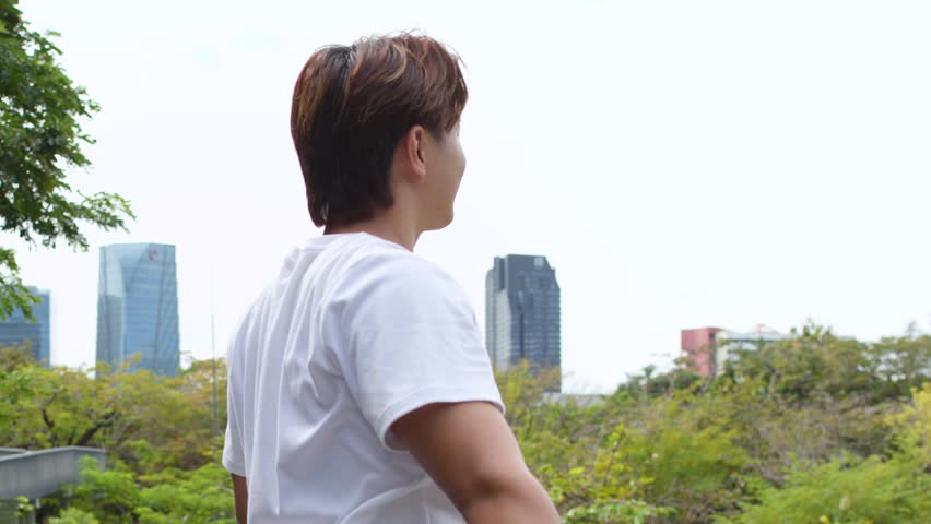 A young Asian man in a white t-shirt stands on a bridge overlooking Bangkok