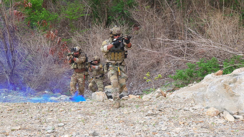 Three soldiers in camouflage tactical gear outdoors on rocky terrain with sparse vegetation. One kneels and aims a rifle forward, while another assists a fallen comrade. Blue smoke in the background suggests a simulated combat or training exercise, highlighting teamwork, readiness, and field operations. Suitable for defense, security, and tactical publications.