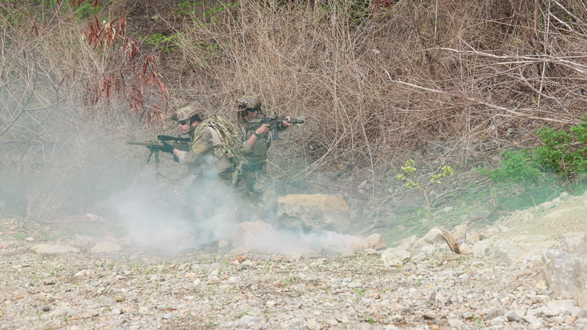 Three soldiers in camouflage tactical gear outdoors on rocky terrain with sparse vegetation. One kneels and aims a rifle forward, while another assists a fallen comrade. Blue smoke in the background suggests a simulated combat or training exercise, highlighting teamwork, readiness, and field operations. Suitable for defense, security, and tactical publications.