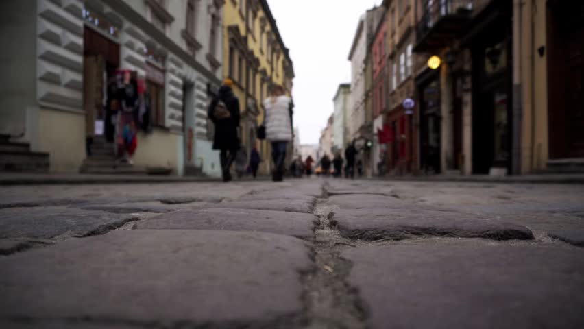 Ground level perspective shows textured cobblestone street surface stretching into distance, framed by old city buildings with architectural detail. Pedestrians walk in background, out of focus, highl