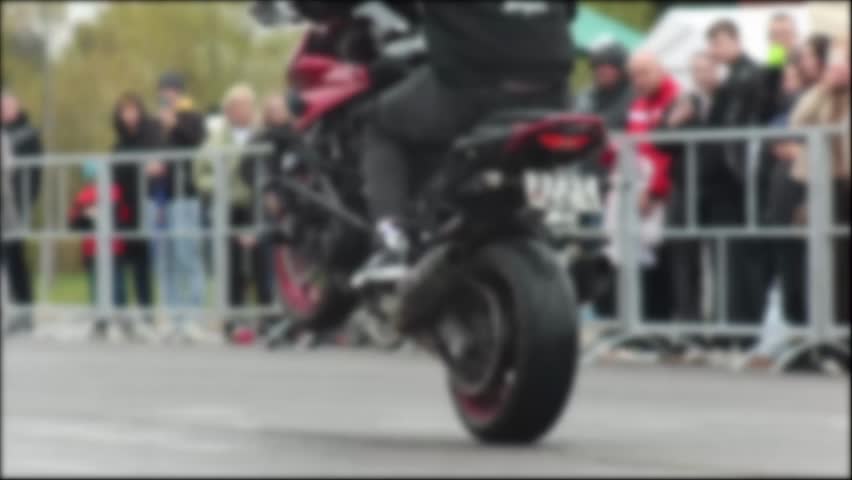 Motorcycle stunt performer executes wheelie trick with precision and control. Audience stands behind barrier, observing thrilling display of skill and courage by rider of sport bike.