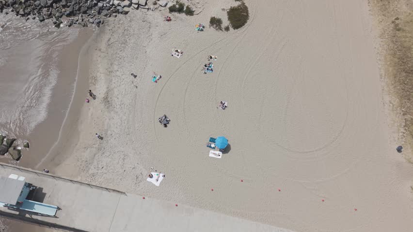 Will Rogers State Beach with beachgoers, blue umbrella and beach towels on sandy shore, gentle waves at water line and rock jetty visible from directly above. California, USA. Aerial top down shot.