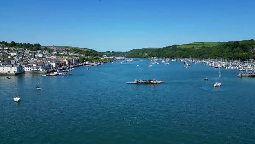 Dartmouth, River Dart, South Devon, England: DRONE VIEWS: The drone flies over the top of the Lower Ferry (vehicle and foot passengers) as it crosses the River Dart heading for Dartmouth. 
Dartmouth is a port town located on the western bank of the River Dart. The Dart is a tidal drowned river (rea) that runs inland as far as Totnes and lies within the South Devon Area of Outstanding Natural Beauty and South Hams district. The town is a popular UK tourist destination renowned both for its scenery and history; the port welcomes occasional cruise liners and has long been associated with its role in seafaring and wars.