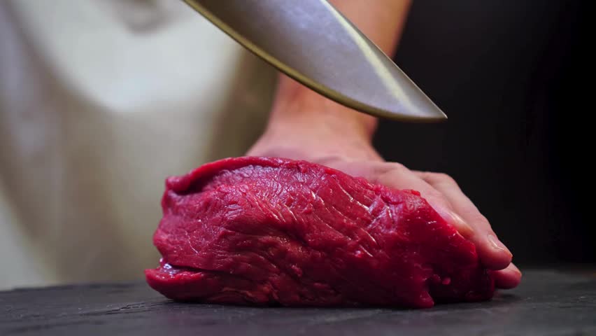 A chef holds a knife carefully, cutting a raw steak lying on a cutting board. Each slice of meat produced is very neat, showing precision in cutting.