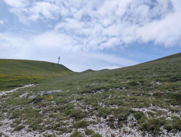 Grassy slope with rocky outcrops and a large cross on the summit, under partly cloudy skies