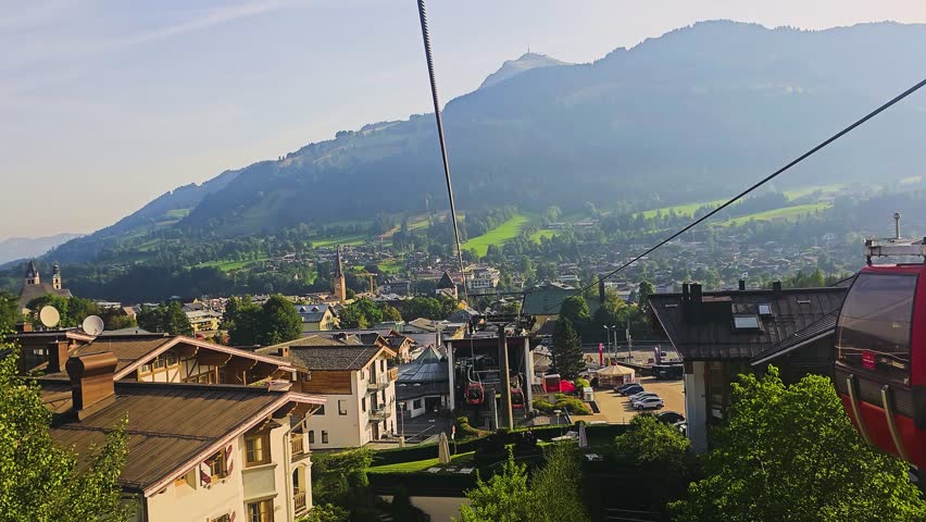 Red gondolas climb the hillside in Kitzbuhel, Austria, surrounded by pine forest, alpine meadows, chalets, and the scenic mountains of the Tyrol summer landscape in summer sunshine.