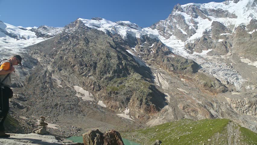 Hiker with backpack admiring glacier lake and Monte Rosa east face from alpine mountain viewpoint on a sunny summer day, trekking adventure in the Italian Alps, Macugnaga