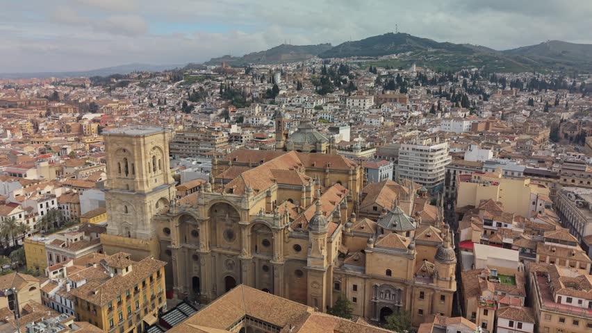 Huge Renaissance Cathedral and Moorish Alhambra complex stand in Granada, Spain. Terracotta roofs fill dense urban cityscape of ancient Andalusian town. 4K aerial of distant Spanish mountains