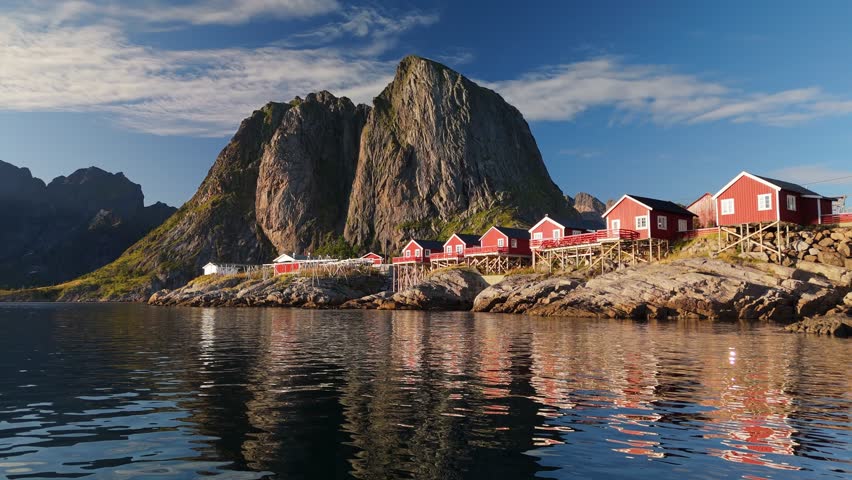 Calm blue fjord water mirrors traditional red fishing cabins of Hamnoy in Lofoten, Norway. Massive rocky mountain peaks tower above historic Norwegian village in this aerial UHD of scenic shore