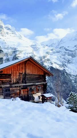 Cozy wooden cabin surrounded by snow in a picturesque mountain landscape. Traditional alpine chalet set against majestic snow-covered peaks under a clear winter sky, creating a peaceful and scenic winter destination.