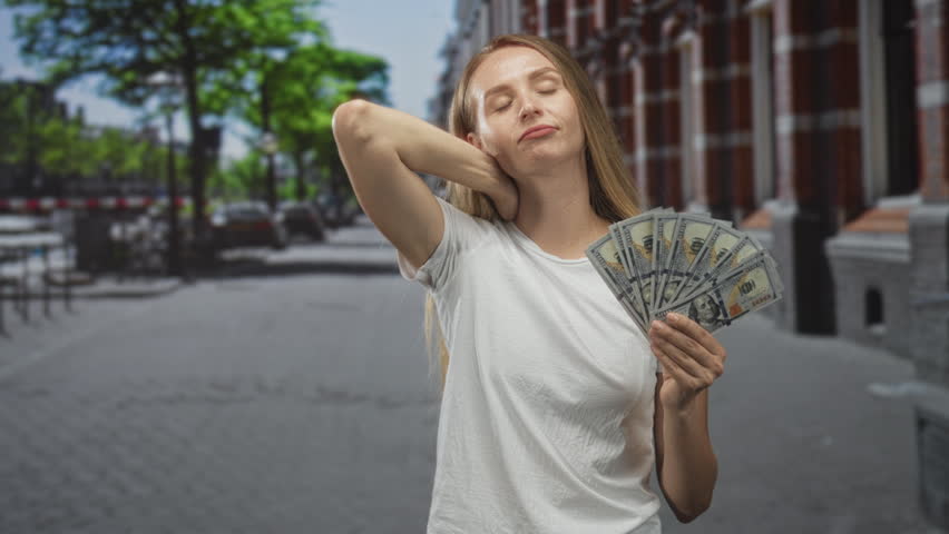 Woman holding fanned dollars, hand to neck on street in white shirt with smirk; confidence abundance.