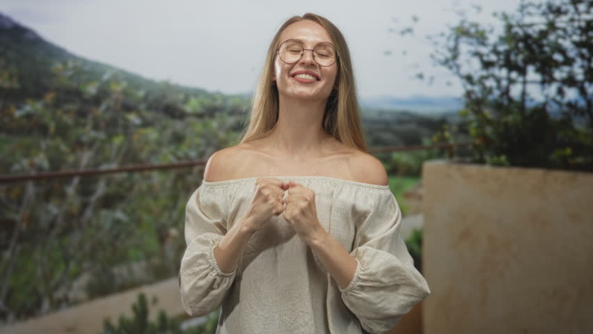 Woman raising fists and smiling on balcony with bare shoulders and glasses, outdoor terrace overlooking hills; joy gratitude hope.