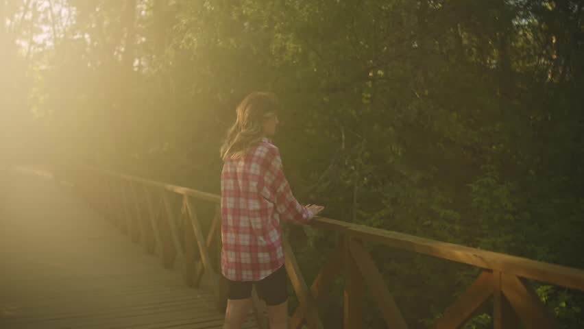 A young woman in a plaid shirt stands on a wooden footbridge and turns back toward the camera, surrounded by dense green trees and warm golden sunlight
