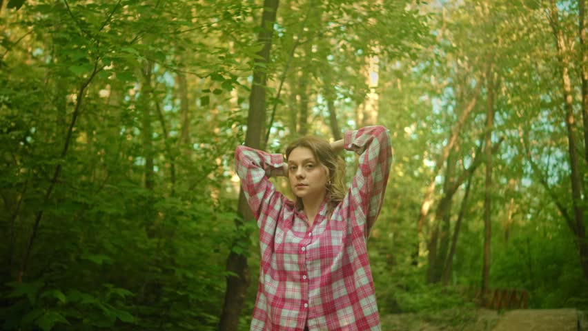 A young woman stands centered among lush trees in soft natural light, posing with raised arms and hands near her hair in a calm woodland setting
