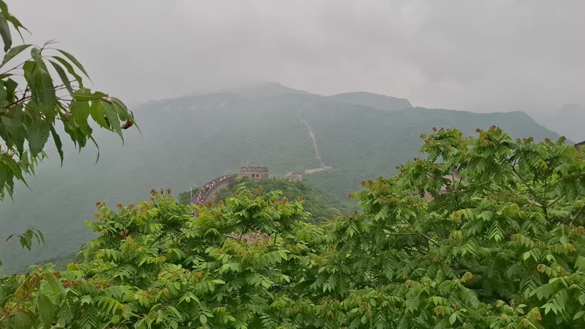 Scenic look at the Great Wall of China at Mutianyu section surrounded by lush green mountains on a cloudy day. Ancient brick fortification framed by vibrant summer tree foliage