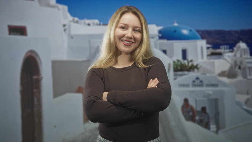 Woman smiling with arms crossed in studio against white greek island buildings and blue dome backdrop; confidence travel.
