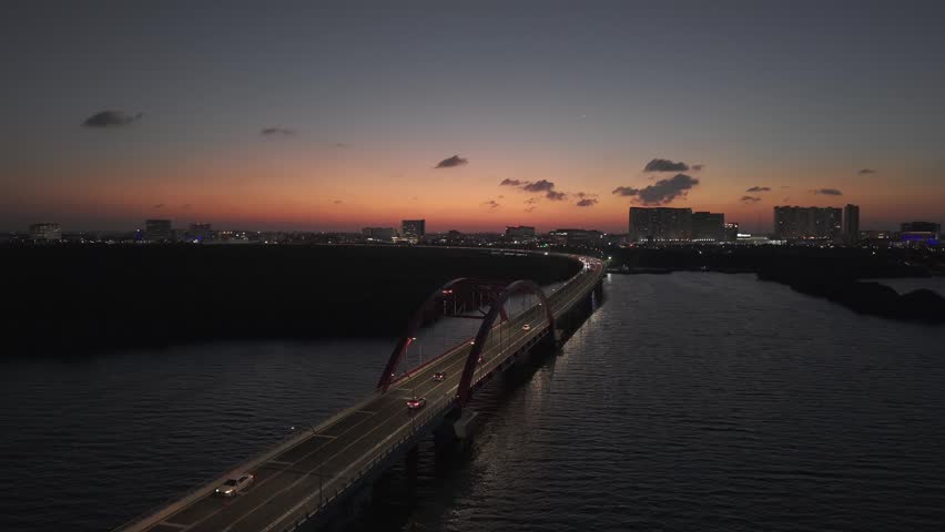 Drone flyover above the Nichupté Bridge at night showcasing illuminated roadways, downtown Cancún lights, and the tropical lagoon below.