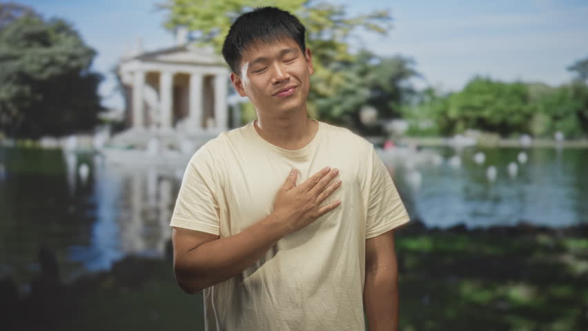 Man points finger at building by park pond in casual tshirt, smiling; confidence relaxed easygoing playful.