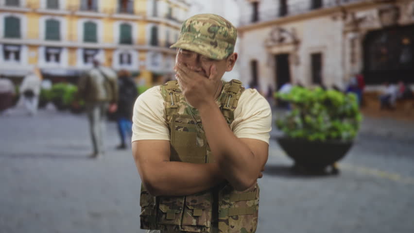 Man soldier in camouflage vest and cap, hand covering mouth in a laughing gesture on a busy street in front of a historic building; pride.
