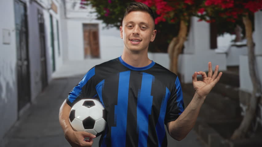 Man in blue and black striped jersey holding soccer ball under arm and making ok sign on narrow whitewashed street; team pride victory.